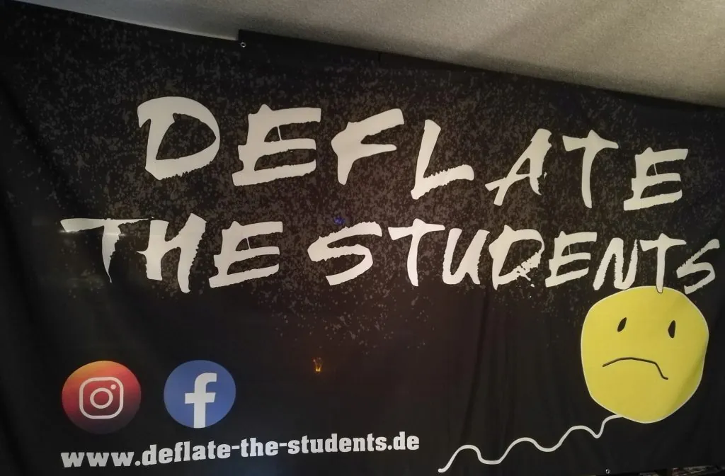 Das schwarze Bandbanner: Schriftzug Deflate The Students, gelber Smiley-Ballon und www.deflate-the-students.de